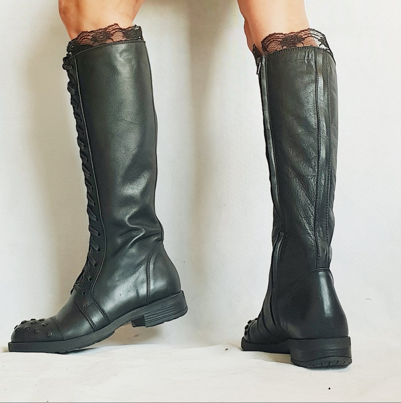 May include: Black knee-high boots with a lace-up front and a zipper on the side. The boots have a square toe and are made of a smooth, dark material. The top of the boots are trimmed with black lace.