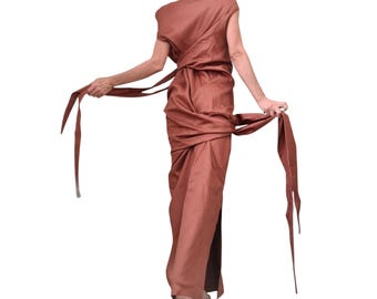 May include: A rust-coloured, draped dress with long, flowing fabric. The dress features a unique wrap design with long, slender ties that extend from the waist. The model is seen from the back, showcasing the dress's elegant silhouette and texture.