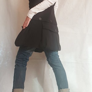 May include: A black vest with a hood and a large pocket on the front. The vest is worn over a white shirt and blue jeans. The person is wearing black boots with zippers on the sides.