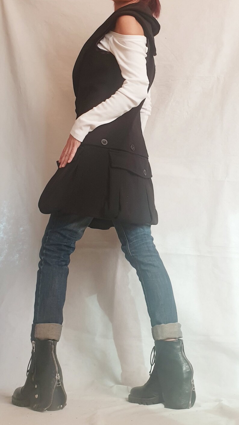 May include: A black vest with a hood and a large pocket on the front. The vest is worn over a white shirt and blue jeans. The person is wearing black boots with zippers on the sides.