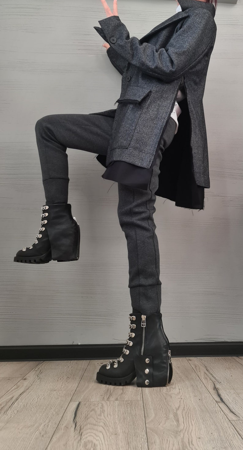 K&ouml;nnte beinhalten: Ein anthrazitfarbener Mantel aus Wollmischgewebe mit Seitenschlitzen, eine dunkelgraue, eng anliegende Hose und schwarze Schn&uuml;rstiefel aus Leder mit silberfarbenen Metallbeschl&auml;gen, einer dicken Profilsohle, einem Rei&szlig;verschluss und drei Nieten.