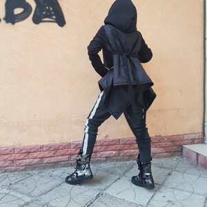 K&ouml;nnte beinhalten: Eine schwarze Kapuzenjacke mit ausgestelltem R&uuml;ckenteil und Bindeband, kombiniert mit einer dunkelgrauen, strukturierten Hose mit hellgrauem Seitenstreifen. Das Outfit wird durch schwarz-graue Stiefel mit Camouflage-Muster abgerundet.