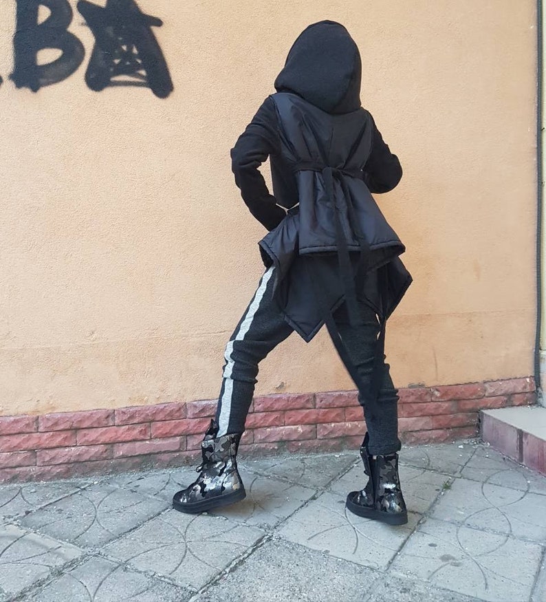 K&ouml;nnte beinhalten: Eine schwarze Kapuzenjacke mit ausgestelltem R&uuml;ckenteil und Bindeband, kombiniert mit einer dunkelgrauen, strukturierten Hose mit hellgrauem Seitenstreifen. Das Outfit wird durch schwarz-graue Stiefel mit Camouflage-Muster abgerundet.