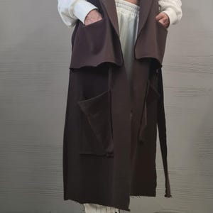 May include: A long, sleeveless dark brown vest featuring a wide collar, two large upper pockets, and two large lower pockets. The vest is worn over a white cropped hooded sweatshirt and matching white sweatpants, paired with black and white sneakers.