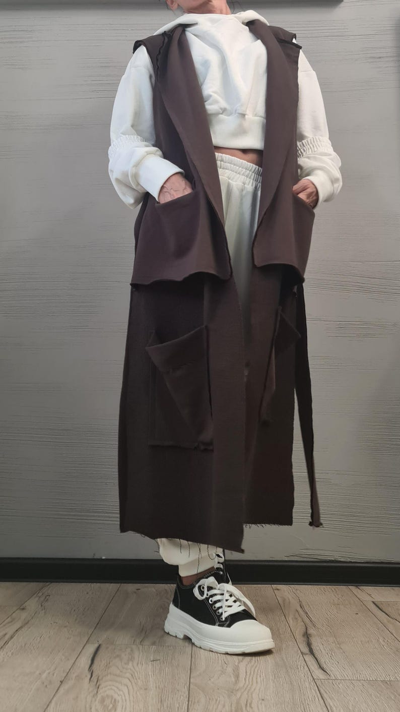 May include: A long, sleeveless dark brown vest featuring a wide collar, two large upper pockets, and two large lower pockets. The vest is worn over a white cropped hooded sweatshirt and matching white sweatpants, paired with black and white sneakers.