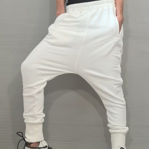 Op de afbeelding: Witte harembroek met een relaxte pasvorm en geribbelde enkels. De broek wordt gecombineerd met een zwarte tanktop met witte bies en wit-zwarte sneakers. De outfit staat tegen een grijze achtergrond.