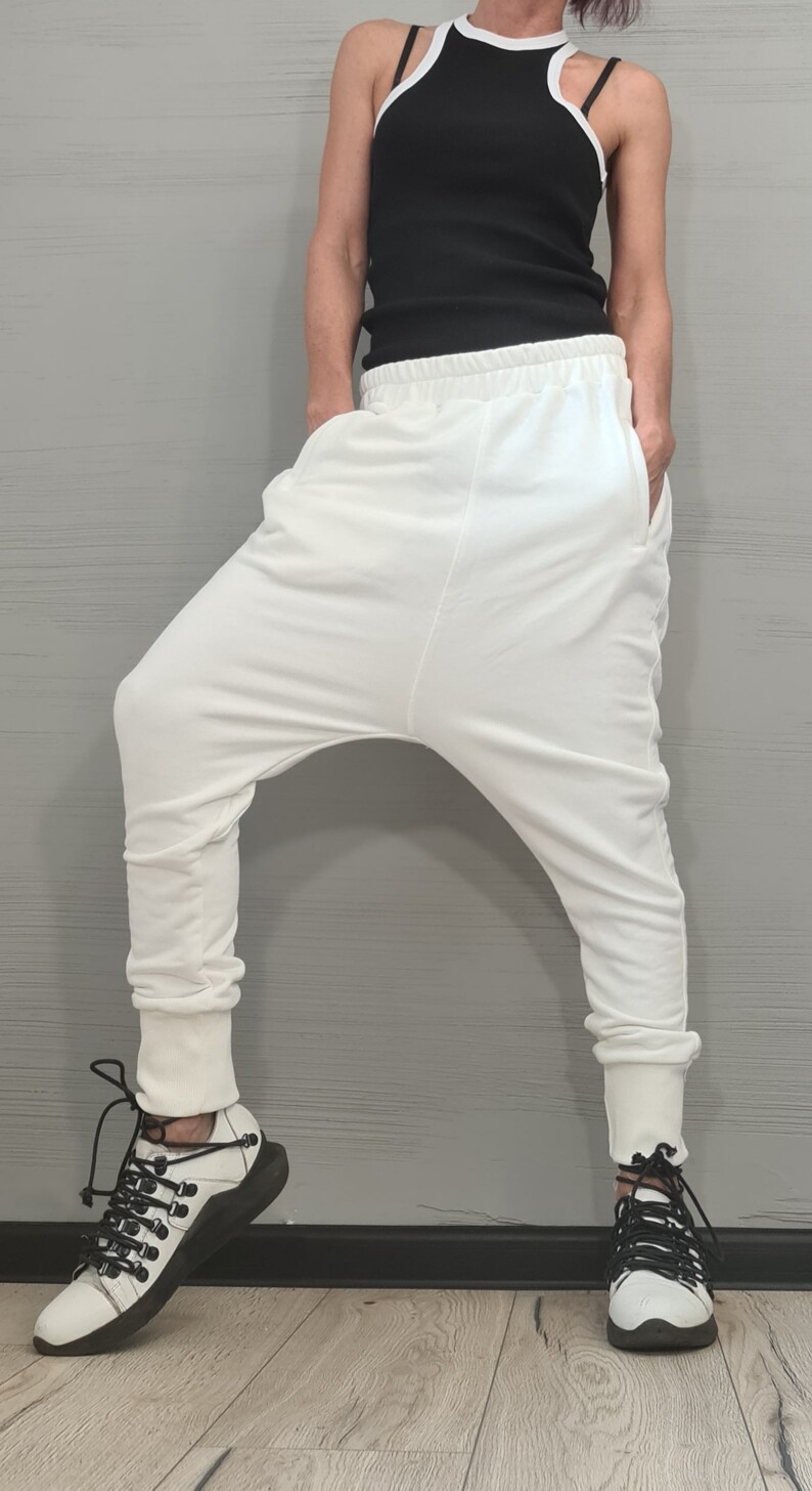 Op de afbeelding: Witte harembroek met een relaxte pasvorm en geribbelde enkels. De broek wordt gecombineerd met een zwarte tanktop met witte bies en wit-zwarte sneakers. De outfit staat tegen een grijze achtergrond.