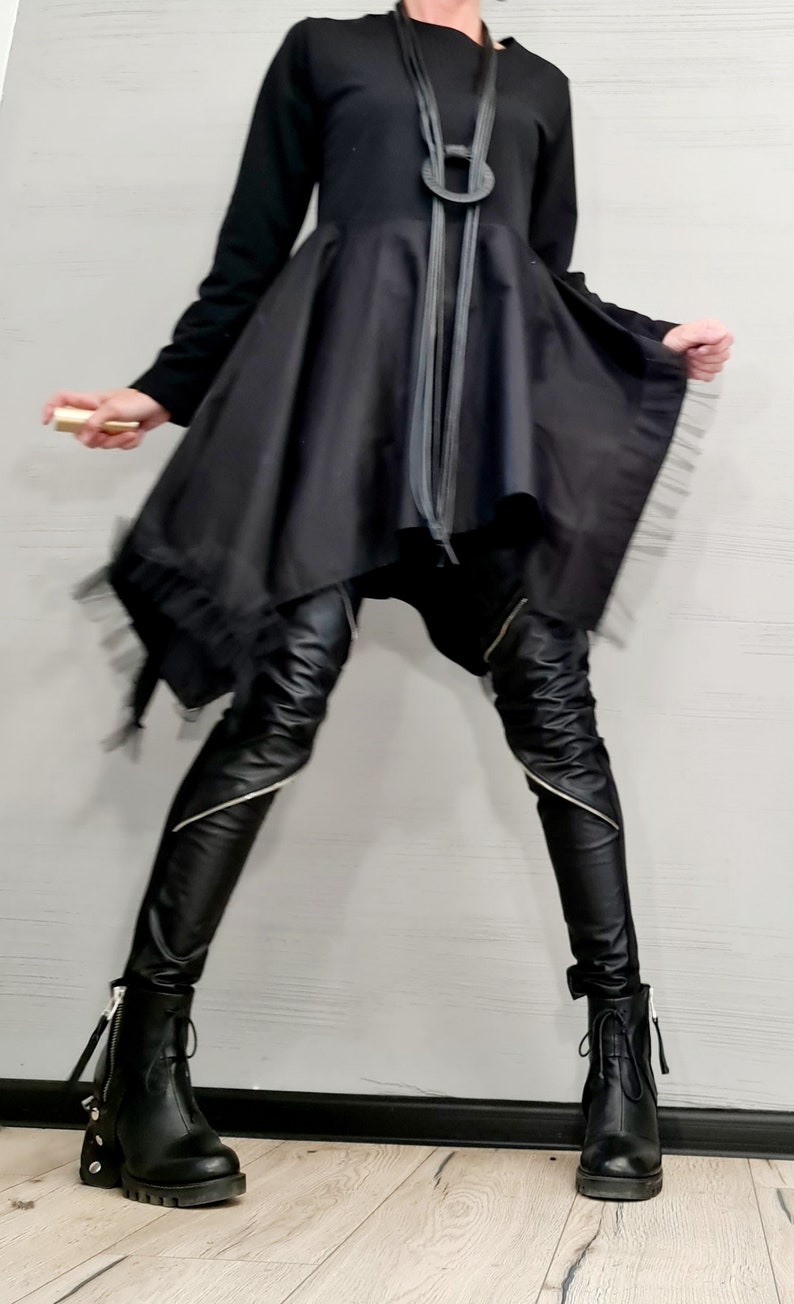 May include: Black outfit featuring a long-sleeved top, asymmetrical skirt, and leather pants with silver zippers. The ensemble includes black boots with chunky heels and a statement necklace. A gold-colored object is held in the hand.
