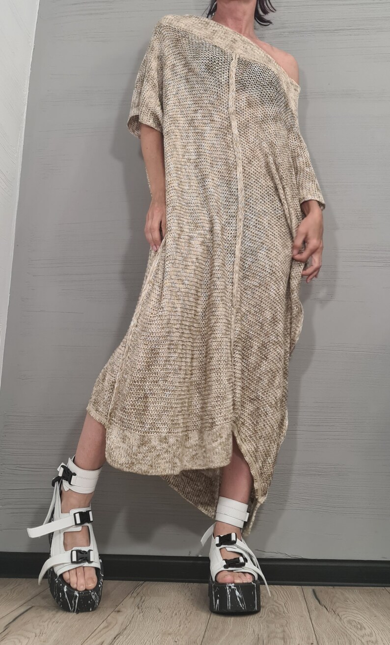 May include: A loose-fitting, beige and white speckled knit dress with short sleeves, an asymmetrical hemline, and a wide neckline. The dress is paired with white platform sandals featuring black buckle straps and a black and white speckled sole.