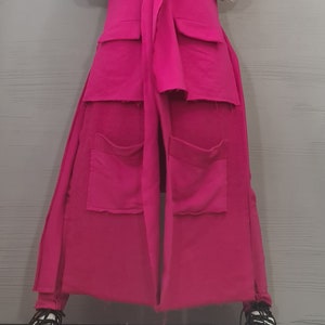May include: A long, sleeveless, hot pink vest featuring a notched lapel, a tie-front closure, and two large patch pockets with raw edges. The vest is worn over a white, long-sleeved, ribbed mock-neck top, paired with white sneakers with black laces.