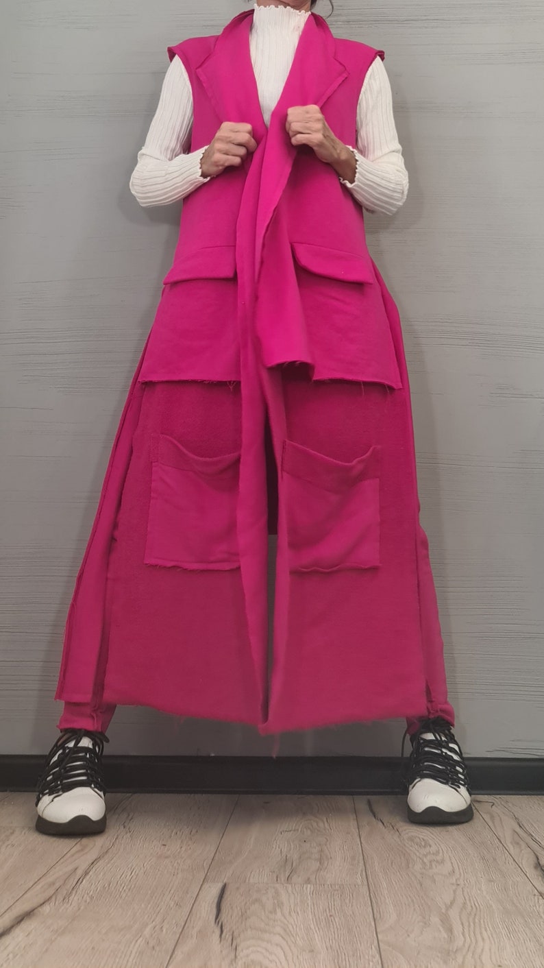 May include: A long, sleeveless, hot pink vest featuring a notched lapel, a tie-front closure, and two large patch pockets with raw edges. The vest is worn over a white, long-sleeved, ribbed mock-neck top, paired with white sneakers with black laces.