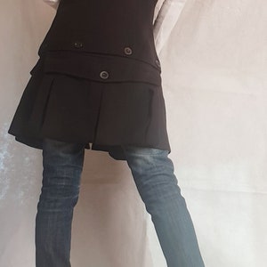 May include: A black long vest with a ruffled hem and a double breasted closure. The vest is worn over a white long-sleeved shirt and blue jeans. The person is wearing black boots with zippers on the sides.