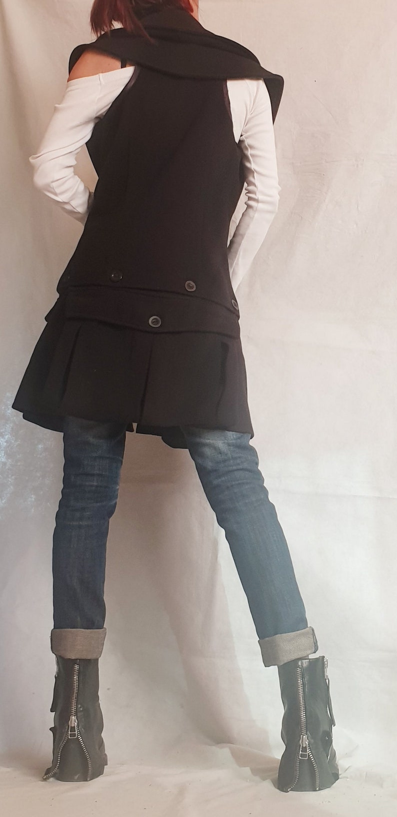 May include: A black long vest with a ruffled hem and a double breasted closure. The vest is worn over a white long-sleeved shirt and blue jeans. The person is wearing black boots with zippers on the sides.