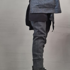 K&ouml;nnte beinhalten: Eine texturierte anthrazitfarbene Jacke mit breitem Kragen und einem Knopfverschluss, getragen &uuml;ber einem schwarzen Oberteil und einer dunkelgrauen, eng anliegenden Hose. Dazu schwarze Lederstiefeletten mit silbernen Rei&szlig;verschl&uuml;ssen und Ketten.