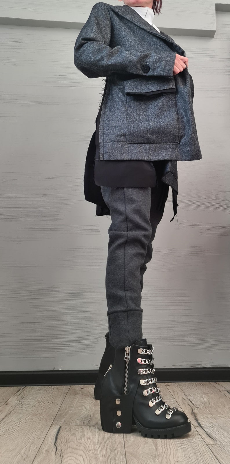 K&ouml;nnte beinhalten: Eine texturierte anthrazitfarbene Jacke mit breitem Kragen und einem Knopfverschluss, getragen &uuml;ber einem schwarzen Oberteil und einer dunkelgrauen, eng anliegenden Hose. Dazu schwarze Lederstiefeletten mit silbernen Rei&szlig;verschl&uuml;ssen und Ketten.