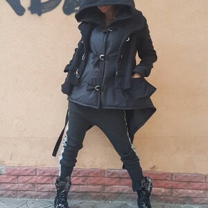 K&ouml;nnte beinhalten: Eine schwarze Kapuzenjacke mit silberfarbenen Metallschnallen &uuml;ber einer schwarzen Steppweste, kombiniert mit einer dunkelgrauen, schmal geschnittenen Hose mit hellgrauen Seitenstreifen und schwarzen, gl&auml;nzenden High-Top-Stiefeln.