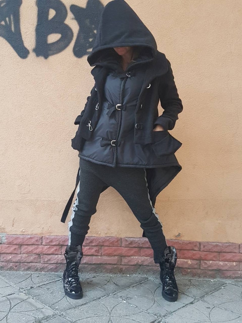 K&ouml;nnte beinhalten: Eine schwarze Kapuzenjacke mit silberfarbenen Metallschnallen &uuml;ber einer schwarzen Steppweste, kombiniert mit einer dunkelgrauen, schmal geschnittenen Hose mit hellgrauen Seitenstreifen und schwarzen, gl&auml;nzenden High-Top-Stiefeln.