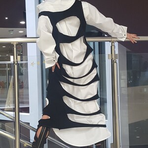 May include: A white button-down shirt is layered under a black, cut-out dress. The dress has a unique design with curved openings. Black sandals with straps complete the outfit. The model is leaning on a metal railing.