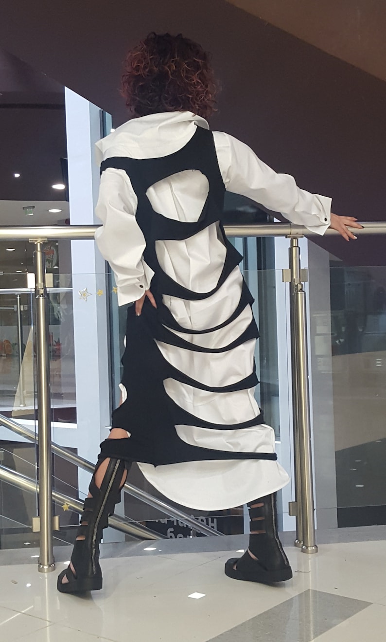 May include: A white button-down shirt is layered under a black, cut-out dress. The dress has a unique design with curved openings. Black sandals with straps complete the outfit. The model is leaning on a metal railing.