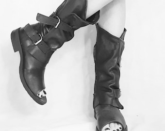 Puede incluir: Un par de botas de cuero negro con hebillas y dedos abiertos. Las botas las lleva una persona con las piernas cruzadas.