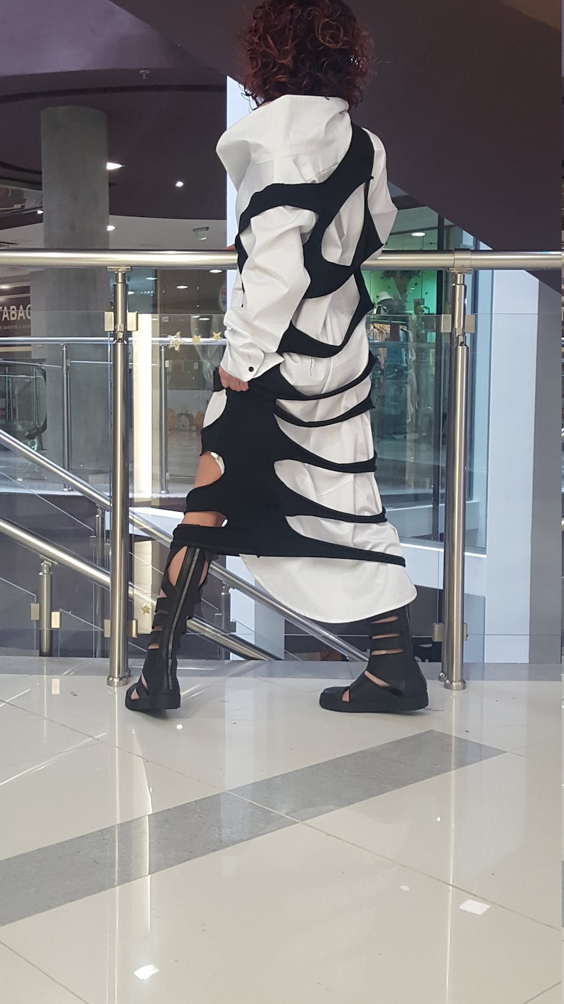 May include: A white shirt and black dress with cut-out details. The dress is layered over the shirt, creating a unique silhouette. Black sandals with multiple straps complete the outfit. The outfit is suitable for a fashion-forward individual.