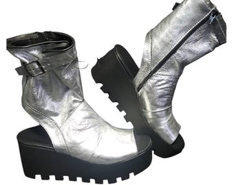 May include: Silver platform sandals with open toes and a chunky black sole. One sandal has a buckle closure, and the other has a zipper. The sandals have a metallic sheen and a unique, edgy design.