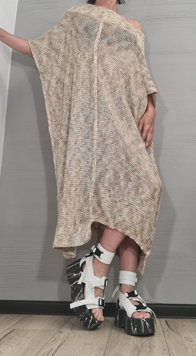 May include: A loose-fitting, beige and white heathered knit tunic dress with a wide neckline and short sleeves. The garment features a vertical center seam and an asymmetrical hemline. It is paired with white platform sandals with black soles.