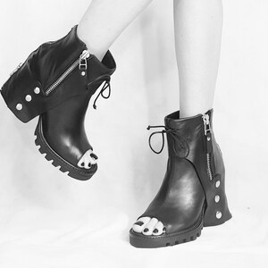 May include: Black leather peep-toe heeled boots with a zipper detail and a chunky heel. The boots have a unique design with a flap that covers the ankle and is secured with a zipper.