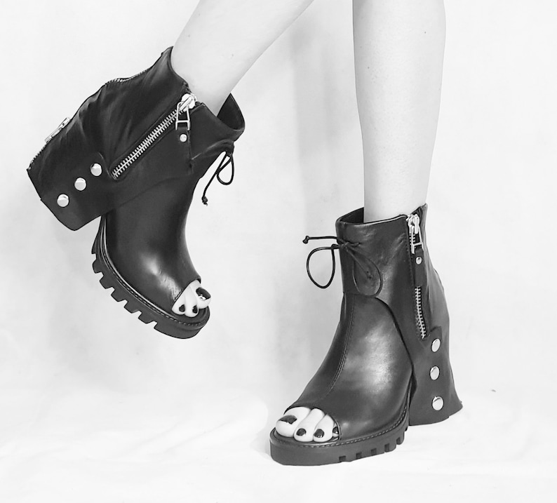 May include: Black leather peep-toe heeled boots with a zipper detail and a chunky heel. The boots have a unique design with a flap that covers the ankle and is secured with a zipper.