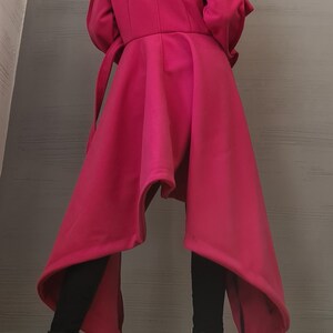 K&ouml;nnte beinhalten: Ein leuchtend pinkfarbener Mantel mit asymmetrischem Saum, langen &Auml;rmeln und einem Bindeg&uuml;rtel. Das Kleidungsst&uuml;ck hat einen strukturierten Kragen und einen ausgestellten Rock, der unter die Knie reicht, kombiniert mit schwarzer Leggings und schwarzen Stiefeletten.
