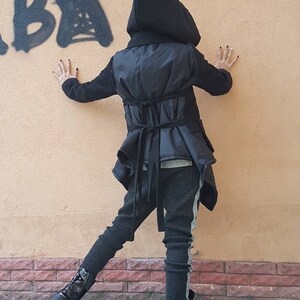 K&ouml;nnte beinhalten: Eine schwarze Kapuzenjacke mit gestepptem R&uuml;ckenteil und zwei horizontalen Bindeb&auml;ndern an der Taille, kombiniert mit einer dunkelgrauen, strukturierten Hose mit hellgrauem Seitenstreifen und schwarzen Schn&uuml;rstiefeletten mit Tarnmuster.