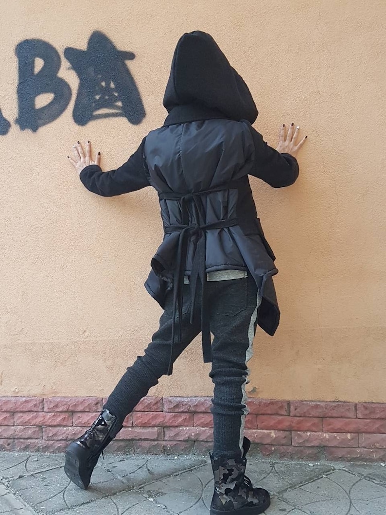K&ouml;nnte beinhalten: Eine schwarze Kapuzenjacke mit gestepptem R&uuml;ckenteil und zwei horizontalen Bindeb&auml;ndern an der Taille, kombiniert mit einer dunkelgrauen, strukturierten Hose mit hellgrauem Seitenstreifen und schwarzen Schn&uuml;rstiefeletten mit Tarnmuster.