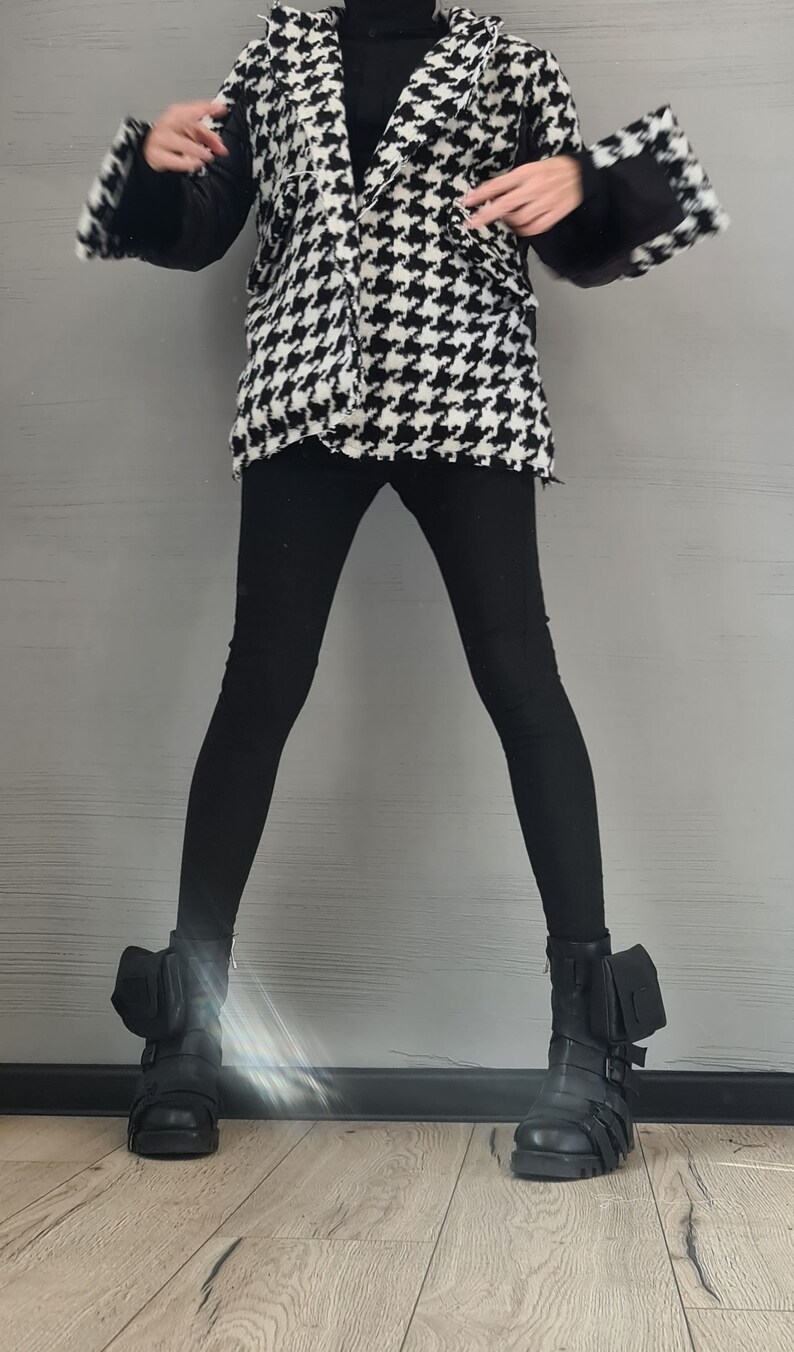 May include: A black and white houndstooth patterned jacket with a notched collar, worn over a black long-sleeved top and black leggings. The outfit is completed with black combat boots featuring side pouches, multiple straps, and buckles.