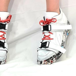 Pu&ograve; includere: Sneakers alte bianche con fibbie nere, lacci color corallo e suola fantasia. Le sneakers hanno cinturini bianchi e un design unico, adatte a persone alla moda.