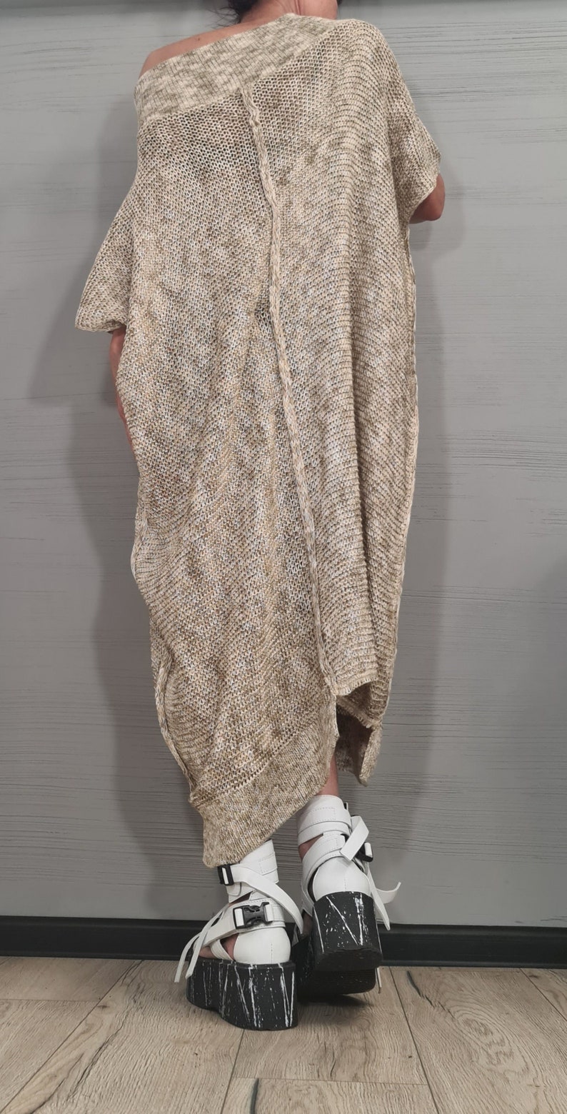 May include: A loose-fitting, beige and white heathered knit tunic with an asymmetrical hemline and a visible vertical seam down the center. The garment is paired with white platform sandals featuring black buckle straps and a black and white speckled sole.