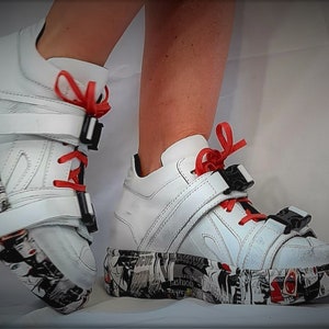 Pu&ograve; includere: Sneakers bianche con plateau, lacci rossi e fibbie nere. Le suole e i lati delle scarpe sono decorate con una stampa grafica in bianco e nero. Le scarpe sono in pelle.