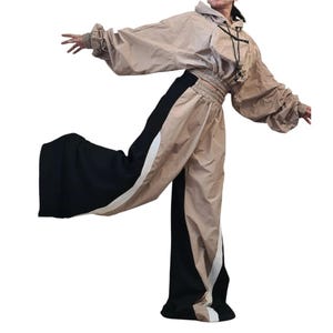 May include: A beige hooded cropped jacket with long sleeves and a front pocket featuring black drawstring cords, paired with wide-leg trousers that have a color-blocked design in beige, black, and white with an elasticized waistband.