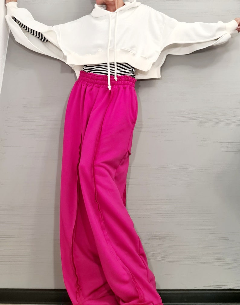 May include: A white cropped hoodie with long sleeves and a drawstring. The sleeves have a black and white striped detail. Paired with bright pink wide-leg sweatpants. A black and white striped shirt is visible underneath the hoodie.