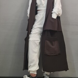 May include: A long, sleeveless dark brown vest featuring large patch pockets, worn over a white hooded sweatshirt and matching white sweatpants. The outfit is completed with black and white sneakers that have thick, chunky white rubber soles.