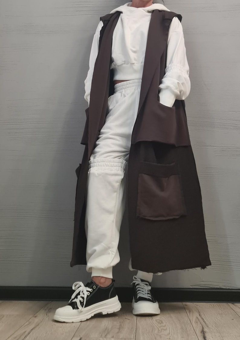 May include: A long, sleeveless dark brown vest featuring large patch pockets, worn over a white hooded sweatshirt and matching white sweatpants. The outfit is completed with black and white sneakers that have thick, chunky white rubber soles.