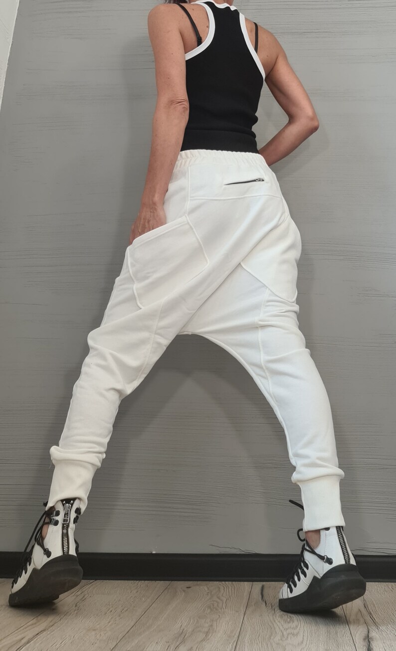 Op de afbeelding: Witte broek met lage kruis, grote zakken en een ritssluiting. De broek wordt gecombineerd met een zwarte tanktop met witte bies en zwart-witte sneakers met ritsen.