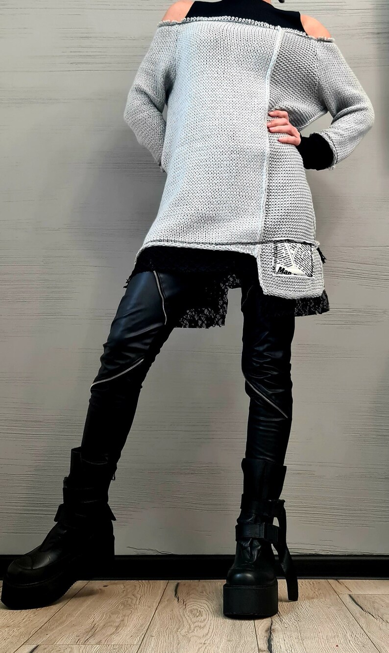 May include: A light gray, long-sleeved, cold-shoulder knit sweater with a raw-edge hem and a small patch featuring newspaper-style text. It is layered over a black lace-trimmed top, black faux leather pants with diagonal zippers, and black platform boots.
