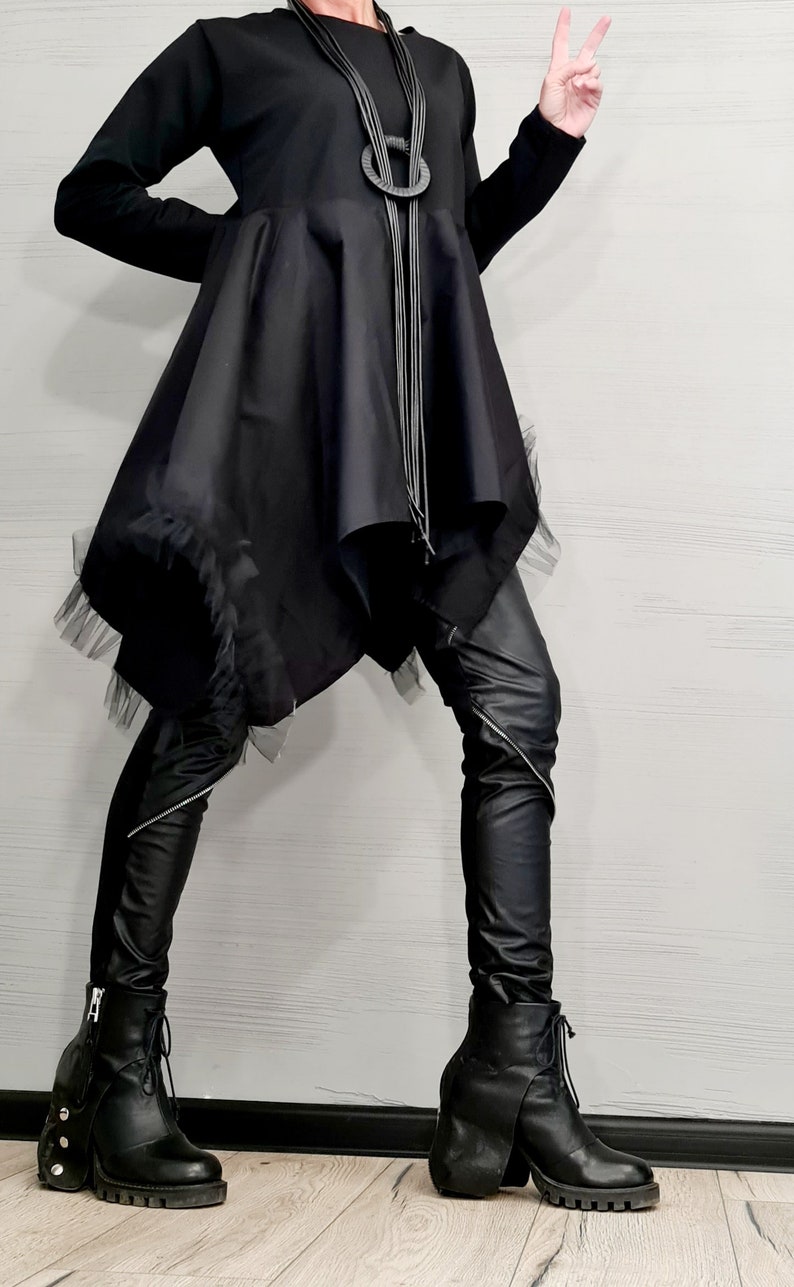 May include: A black outfit featuring a long-sleeved top with an asymmetrical skirt and black leather pants. The outfit is accessorized with a layered necklace and chunky black boots with zippers and laces. The overall style is modern and edgy.