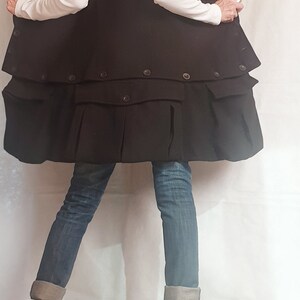 May include: A black, oversized, double-breasted coat with a high collar and pockets. The coat is worn over a white long-sleeved shirt and blue jeans. The person is wearing black boots with zippers on the sides.