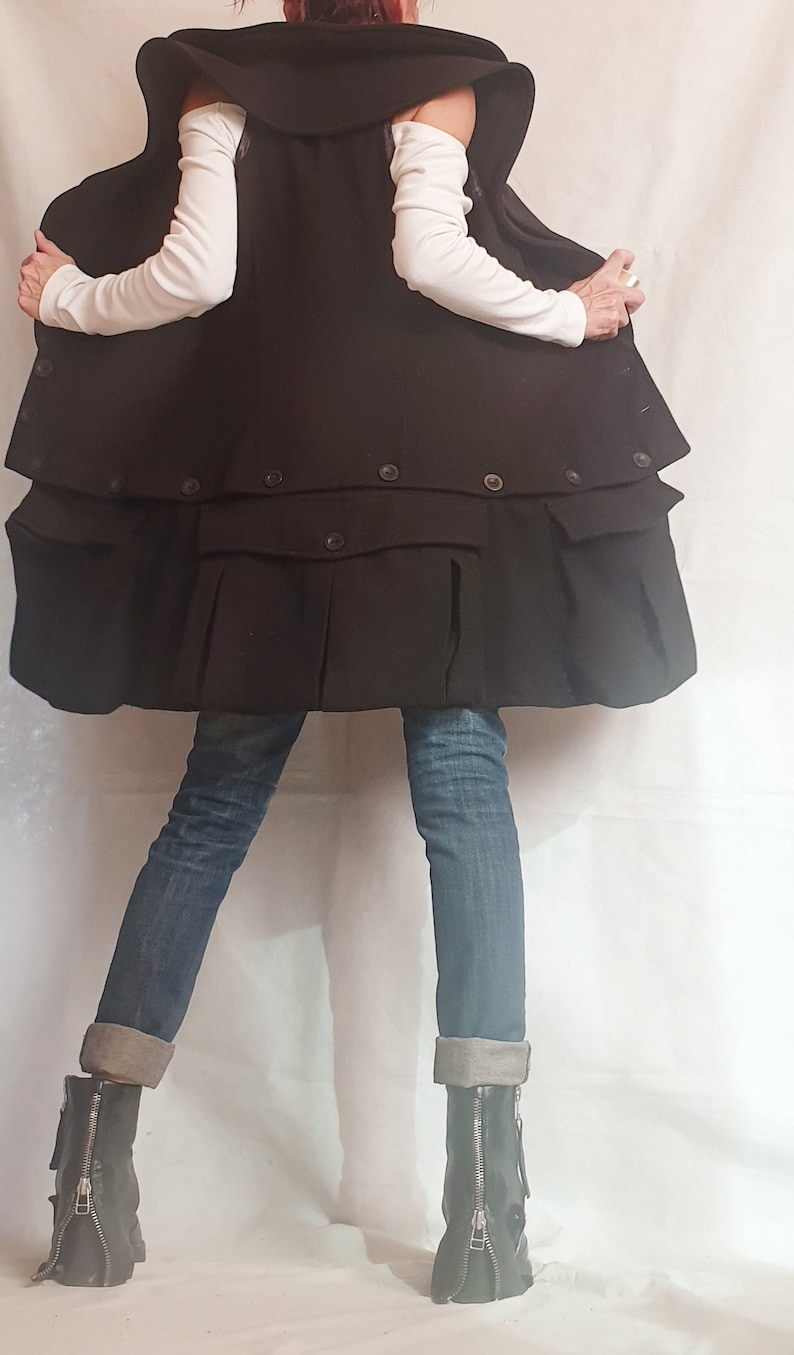 May include: A black, oversized, double-breasted coat with a high collar and pockets. The coat is worn over a white long-sleeved shirt and blue jeans. The person is wearing black boots with zippers on the sides.