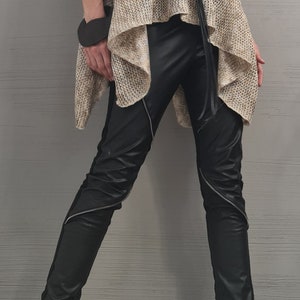 May include: A sleeveless beige knit turtleneck tunic with an asymmetrical hem, paired with black faux leather pants featuring diagonal silver zippers. A long black cord necklace with a circular pendant and black buckled ankle boots complete the look.