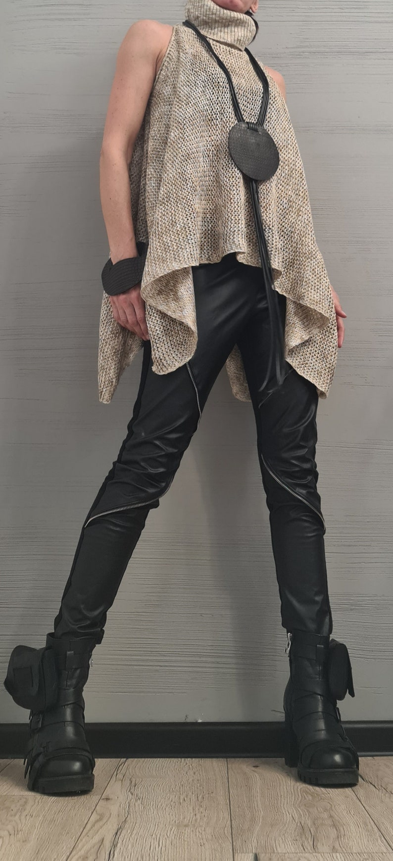 May include: A sleeveless beige knit turtleneck tunic with an asymmetrical hem, paired with black faux leather pants featuring diagonal silver zippers. A long black cord necklace with a circular pendant and black buckled ankle boots complete the look.