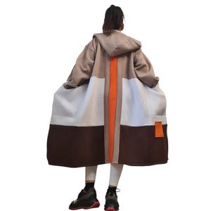 Puede incluir: Un abrigo con capucha con un diseño de bloques de color. El abrigo presenta una combinación de colores marrón, beige, blanco y naranja. Tiene una franja naranja vertical en la espalda y bolsillos grandes. La persona lleva leggings blancos y zapatillas negras.
