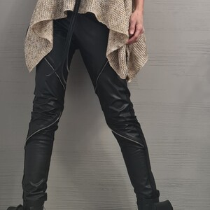 May include: A beige sleeveless turtleneck top with a loose knit texture, paired with black trousers featuring diagonal silver zippers on the thighs. A black circular pendant hangs from a long cord. Black boots with side pouches and buckles are worn.