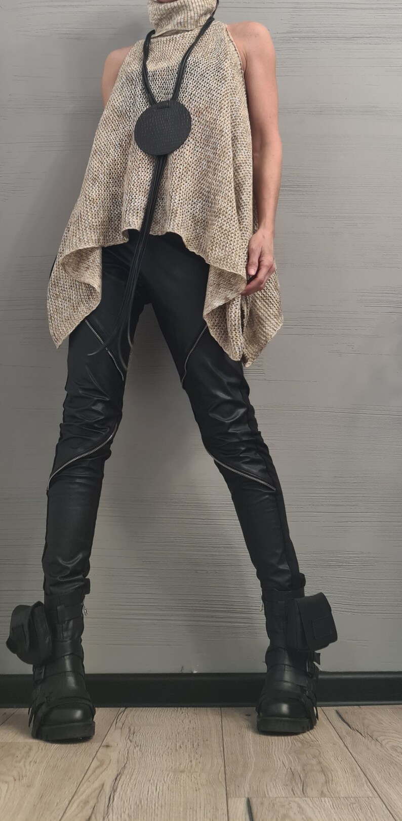 May include: A beige sleeveless turtleneck top with a loose knit texture, paired with black trousers featuring diagonal silver zippers on the thighs. A black circular pendant hangs from a long cord. Black boots with side pouches and buckles are worn.